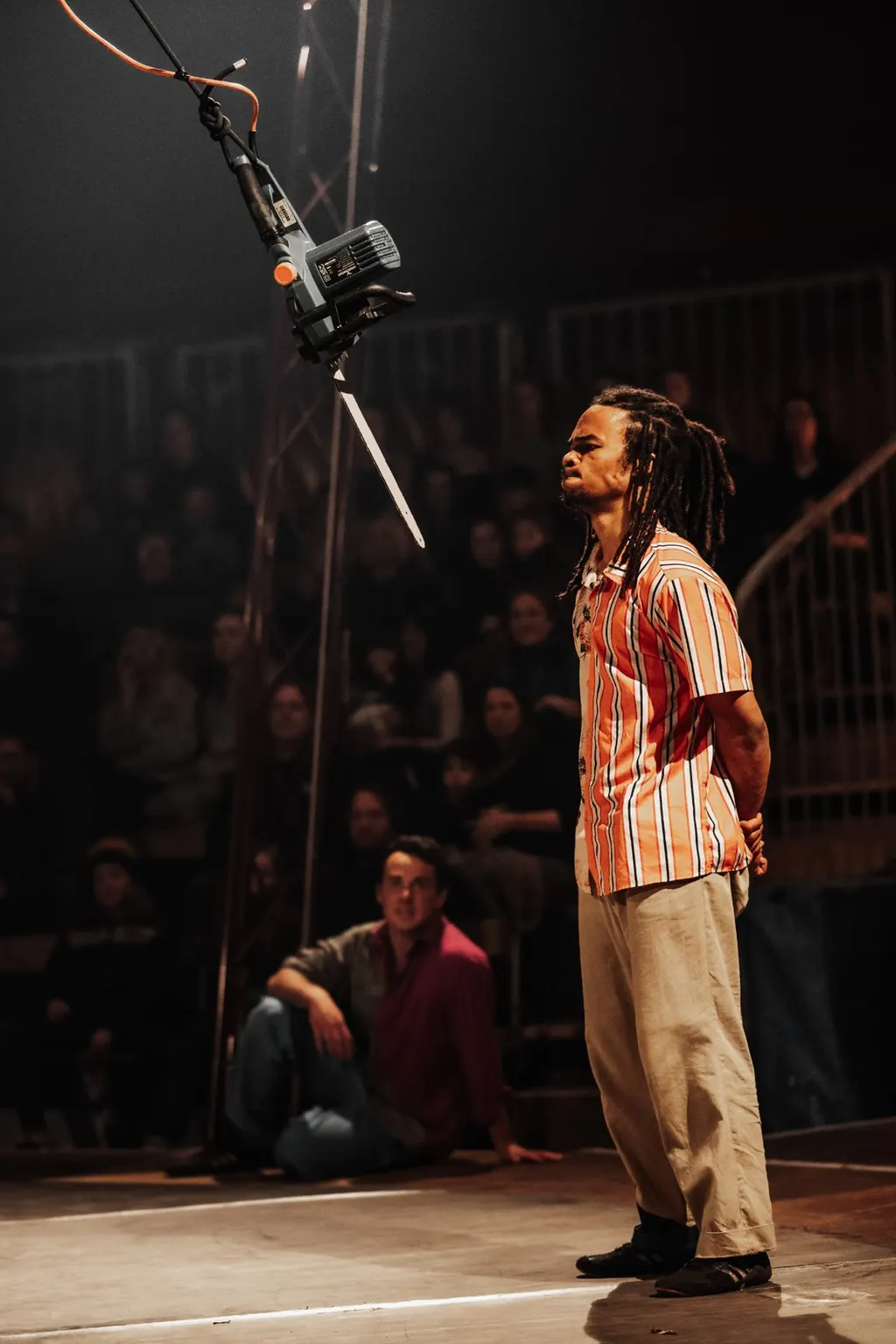 A performer stands beneath a swinging chainsaw in The Wing-Scuffle Spectacular, the audience watching in the round