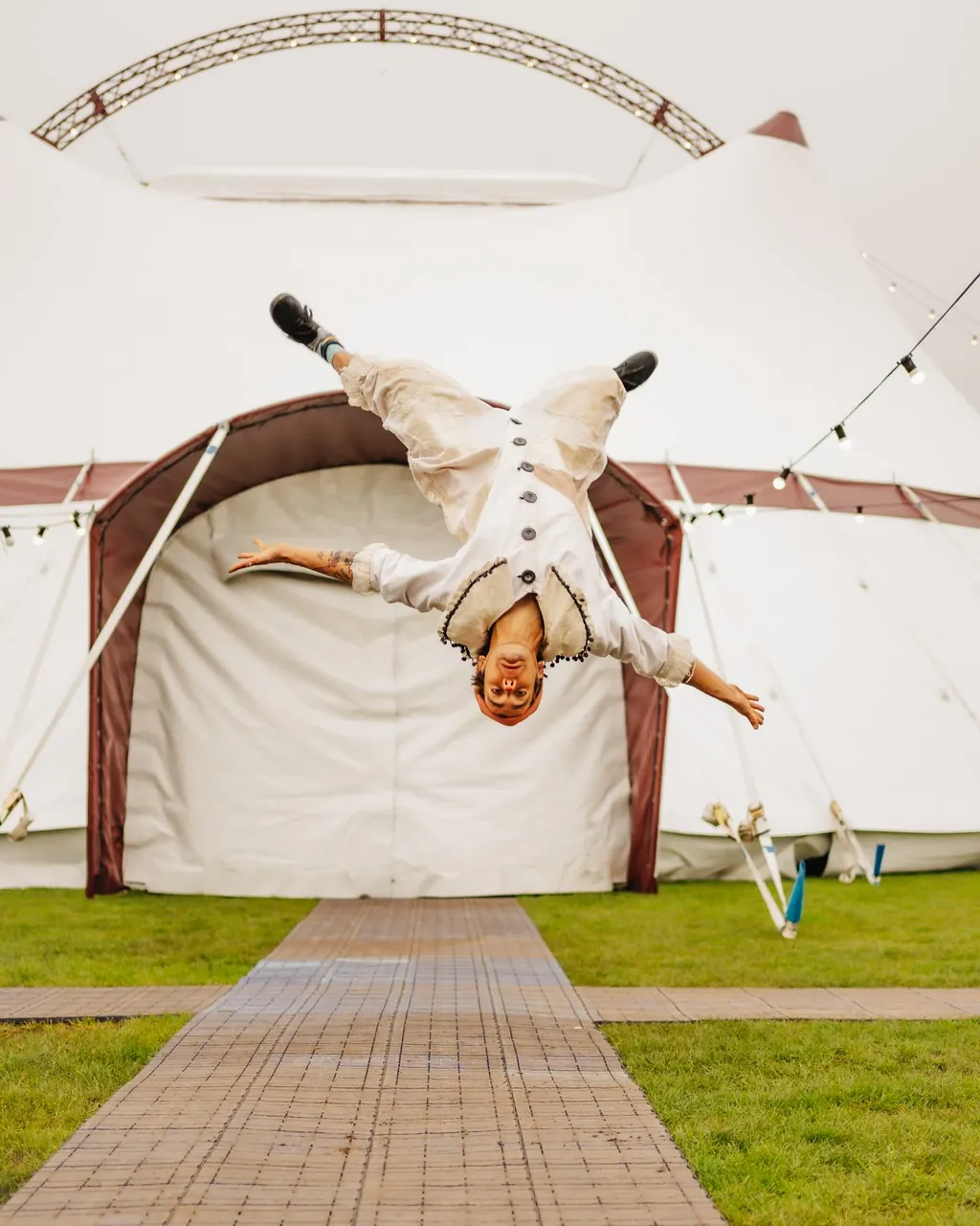 A performer in white flips upside down on the path outside the Revel Puck big top